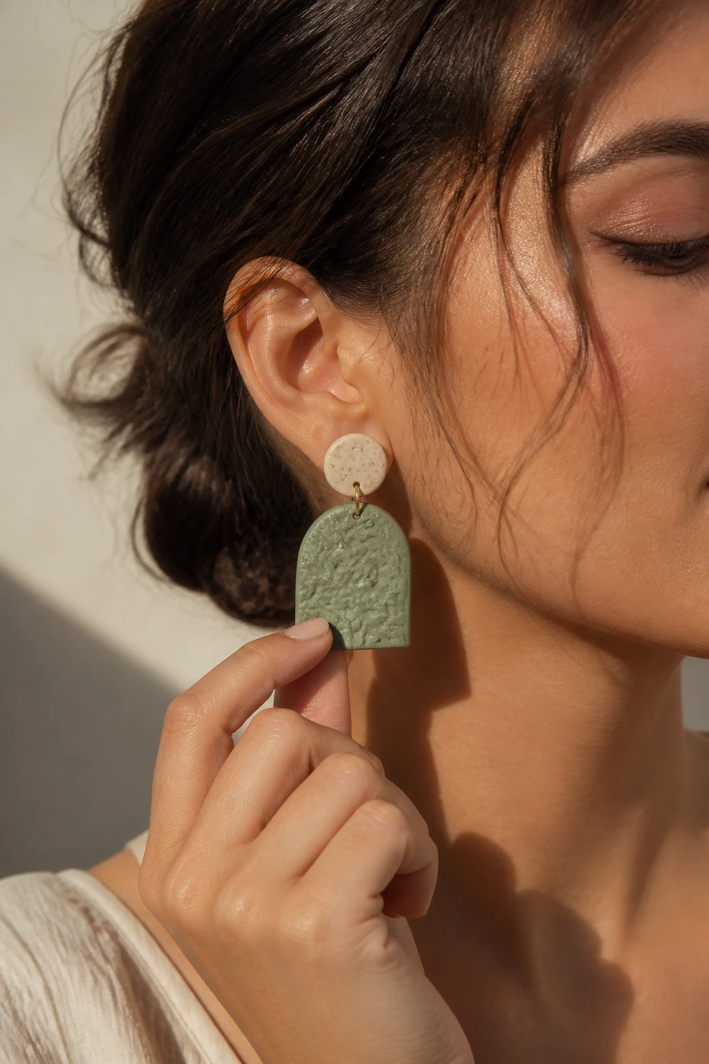 A product-in-use scene showing a hand modeling a pair of weighty yet lightweight polymer clay earrings, with a focus on the ear area, subtle jewelry photography lighting, and a soft shadow to convey dimension.